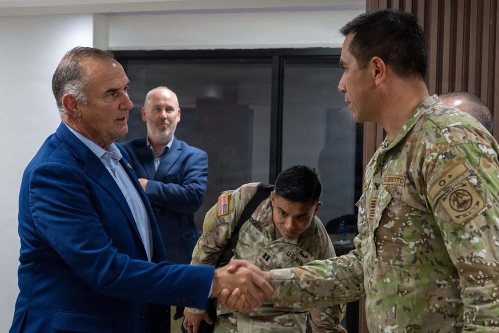 A civilian in a blue blazer is shaking hands with a military officer wearing camouflage uniform with 'EJERCITO' visible on the patch, suggesting a Latin American army affiliation. In the background, a U.S. Army soldier (with 'U.S. ARMY' and 'ROGERS' name tape visible) is bent over, and another civilian in a blue jacket observes the interaction. The setting appears to be an indoor meeting or briefing room.