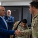 A civilian in a blue blazer is shaking hands with a military officer wearing camouflage uniform with 'EJERCITO' visible on the patch, suggesting a Latin American army affiliation. In the background, a U.S. Army soldier (with 'U.S. ARMY' and 'ROGERS' name tape visible) is bent over, and another civilian in a blue jacket observes the interaction. The setting appears to be an indoor meeting or briefing room.