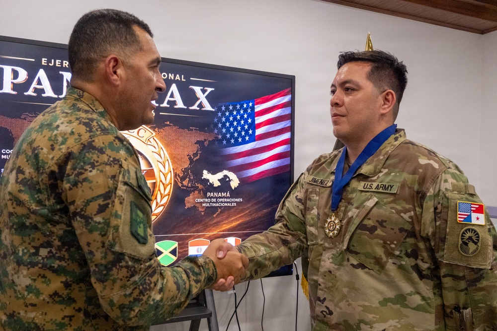 A U.S. Army soldier identified by the name tape 'JIMENEZ' is receiving a medal during what appears to be a multinational military ceremony. He is shaking hands with a soldier wearing a different camouflage pattern, likely from a Latin American military. The event appears connected to 'PANAMAX,' a multinational military exercise centered in Panama.