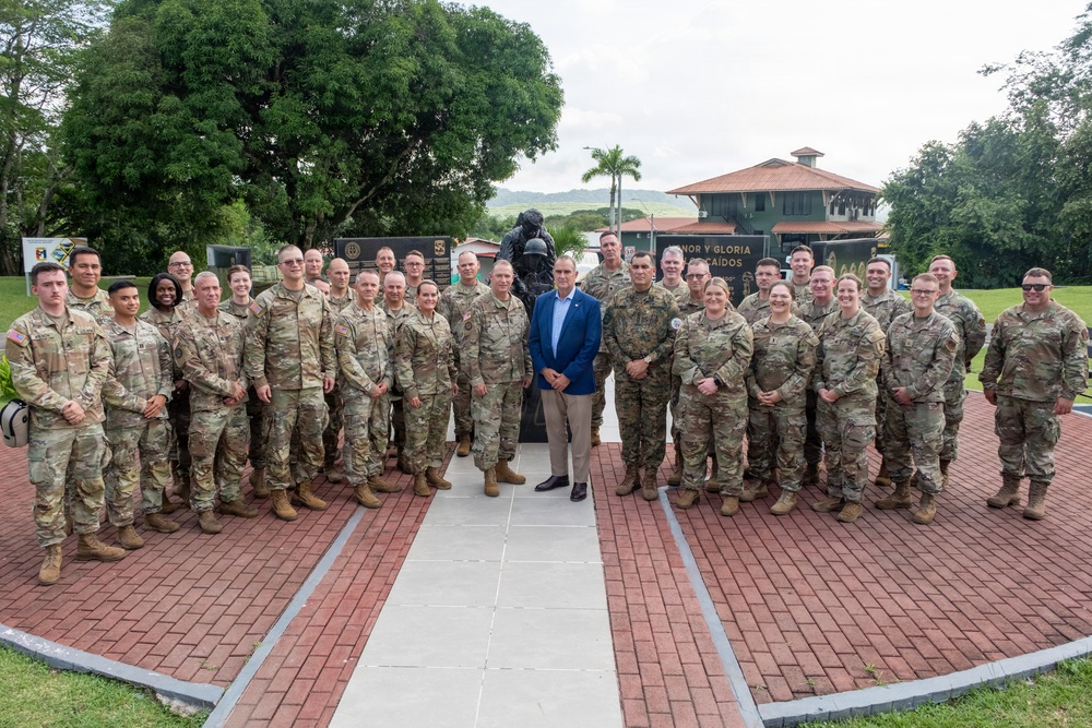 A large group of approximately 30 military personnel in U.S. Army camouflage uniforms pose for a formal group photograph alongside one civilian in a blue suit. They are gathered in front of a bronze memorial statue on a brick plaza, with a sign partially reading 'HONOR Y GLORIA' and 'CAIDOS' visible in the background, suggesting a fallen soldiers memorial. The setting appears to be a military installation in a tropical or subtropical location.