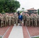 A large group of approximately 30 military personnel in U.S. Army camouflage uniforms pose for a formal group photograph alongside one civilian in a blue suit. They are gathered in front of a bronze memorial statue on a brick plaza, with a sign partially reading 'HONOR Y GLORIA' and 'CAIDOS' visible in the background, suggesting a fallen soldiers memorial. The setting appears to be a military installation in a tropical or subtropical location.