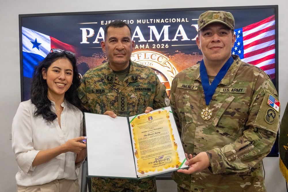 Three individuals pose for a formal military ceremony photo in front of a screen displaying 'Ejercicio Multinacional PANAMAX Panamá 2026' with Panamanian and American flags. A U.S. Army soldier identified by nametape as 'JIMENEZ' is being awarded a medal and certificate, alongside a Panamanian military officer identified as 'L. SOLIS' from SENAFRONT. The event appears to be a bilateral military cooperation recognition ceremony.