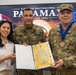 Three individuals pose for a formal military ceremony photo in front of a screen displaying 'Ejercicio Multinacional PANAMAX Panamá 2026' with Panamanian and American flags. A U.S. Army soldier identified by nametape as 'JIMENEZ' is being awarded a medal and certificate, alongside a Panamanian military officer identified as 'L. SOLIS' from SENAFRONT. The event appears to be a bilateral military cooperation recognition ceremony.