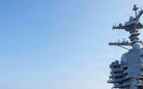 A large group of U.S. Navy personnel are conducting a Foreign Object Debris (FOD) walkdown on the flight deck of an aircraft carrier. Crew members wearing color-coded jerseys — yellow, green, and blue — are walking in a line formation across the deck, searching for debris. The ship's island superstructure is visible on the right side, flying an American flag.