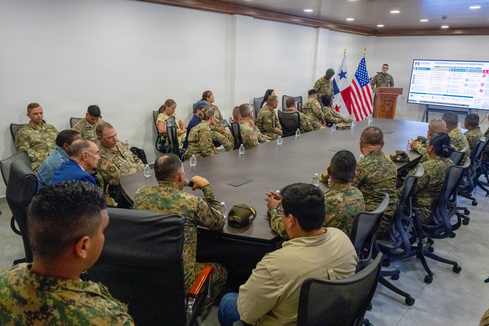 A multinational military briefing is taking place in a conference room, with personnel from at least two nations seated around a large oval table. A presenter stands at a podium beside a display screen showing a slide titled 'Requerimientos Estratégicos Prioritarios' (Priority Strategic Requirements). Both the United States flag and what appears to be the Panamanian flag are displayed prominently at the front of the room.