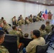 A multinational military briefing is taking place in a conference room, with personnel from at least two nations seated around a large oval table. A presenter stands at a podium beside a display screen showing a slide titled 'Requerimientos Estratégicos Prioritarios' (Priority Strategic Requirements). Both the United States flag and what appears to be the Panamanian flag are displayed prominently at the front of the room.