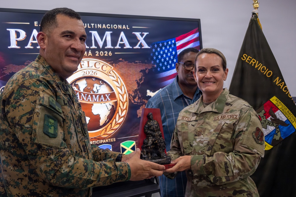 Two military personnel are exchanging a commemorative figurine trophy during what appears to be an official ceremony related to the PANAMAX 2026 multinational exercise. A U.S. Air Force member (name tag reads 'SETTLE') is receiving or presenting the award alongside a soldier in camouflage uniform, with a third individual visible in the background. The setting includes a PANAMAX 2026 FLCC exercise banner and a Panamanian 'Servicio Nacional de Fronteras' flag.