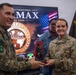 Two military personnel are exchanging a commemorative figurine trophy during what appears to be an official ceremony related to the PANAMAX 2026 multinational exercise. A U.S. Air Force member (name tag reads 'SETTLE') is receiving or presenting the award alongside a soldier in camouflage uniform, with a third individual visible in the background. The setting includes a PANAMAX 2026 FLCC exercise banner and a Panamanian 'Servicio Nacional de Fronteras' flag.