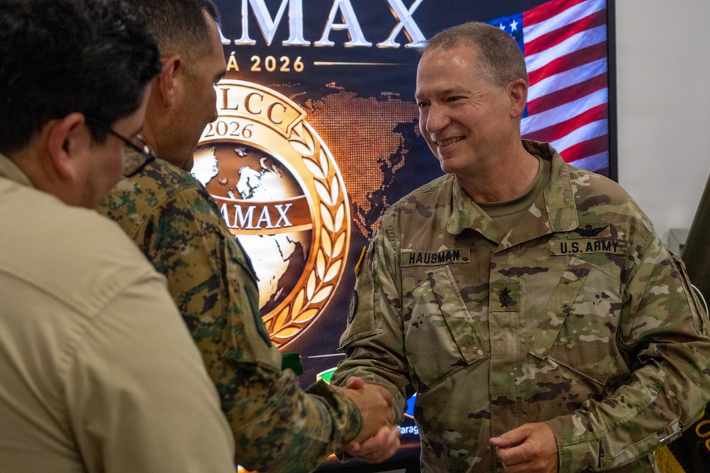 A U.S. Army soldier named Hausman, wearing OCP (Operational Camouflage Pattern) uniform, is shaking hands with another military personnel at what appears to be an AMAX/LCCAMAX 2026 military event or exercise. The background displays an official event logo featuring a world map medallion design and an American flag. The setting suggests a multinational military conference or exercise coordination meeting.
