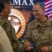 A U.S. Army soldier named Hausman, wearing OCP (Operational Camouflage Pattern) uniform, is shaking hands with another military personnel at what appears to be an AMAX/LCCAMAX 2026 military event or exercise. The background displays an official event logo featuring a world map medallion design and an American flag. The setting suggests a multinational military conference or exercise coordination meeting.