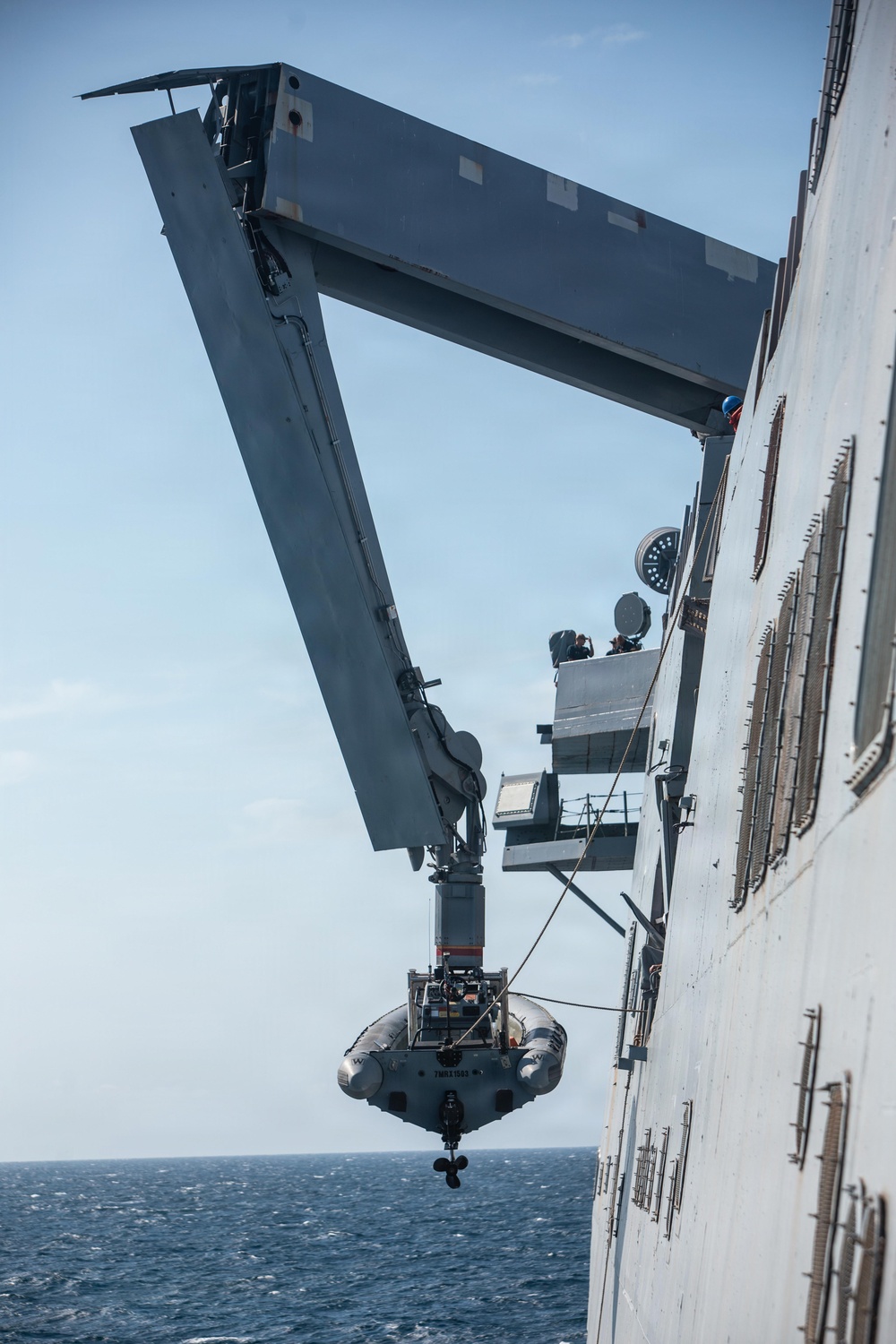 A rigid inflatable boat (RIB) is being deployed or recovered via a large davit crane system mounted on the side of a naval warship at sea. The crane arm extends outward over the water, suspending the small boat alongside the ship's hull. At least one crew member in a blue helmet is visible on the ship's superstructure overseeing the operation.