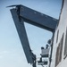 A rigid inflatable boat (RIB) is being deployed or recovered via a large davit crane system mounted on the side of a naval warship at sea. The crane arm extends outward over the water, suspending the small boat alongside the ship's hull. At least one crew member in a blue helmet is visible on the ship's superstructure overseeing the operation.