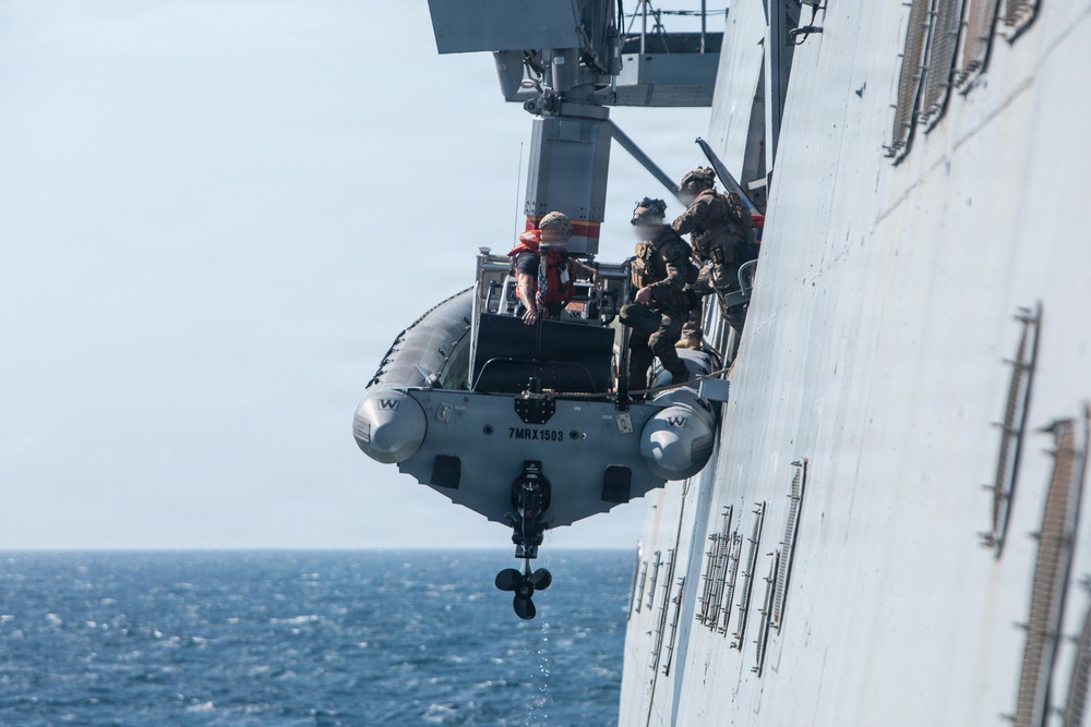 A rigid-hull inflatable boat (RHIB) is being deployed or recovered via davit crane on the side of a large naval vessel at sea. Several personnel in military tactical gear are visible on or near the boat during the operation. The image appears to document a maritime boarding or launch/recovery evolution.