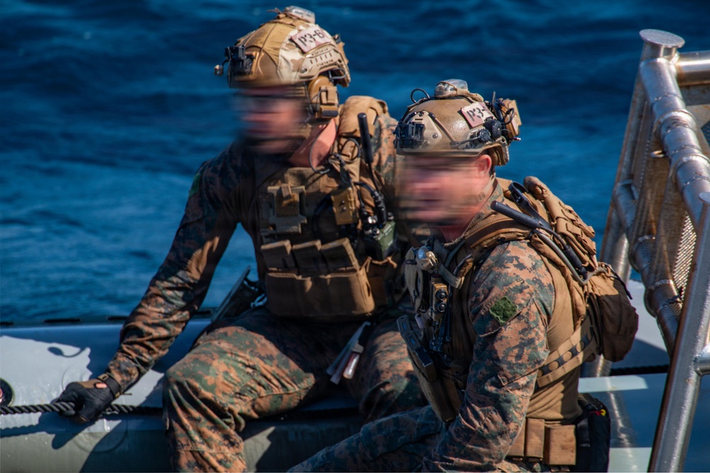 Two military personnel in full tactical gear are seated on what appears to be a rigid inflatable boat (RIB) on open water. Both individuals are wearing MARPAT (Marine pattern) camouflage uniforms with plate carriers, tactical helmets, and communications equipment. Their faces appear motion-blurred, likely intentionally obscured for operational security purposes.