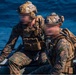 Two military personnel in full tactical gear are seated on what appears to be a rigid inflatable boat (RIB) on open water. Both individuals are wearing MARPAT (Marine pattern) camouflage uniforms with plate carriers, tactical helmets, and communications equipment. Their faces appear motion-blurred, likely intentionally obscured for operational security purposes.