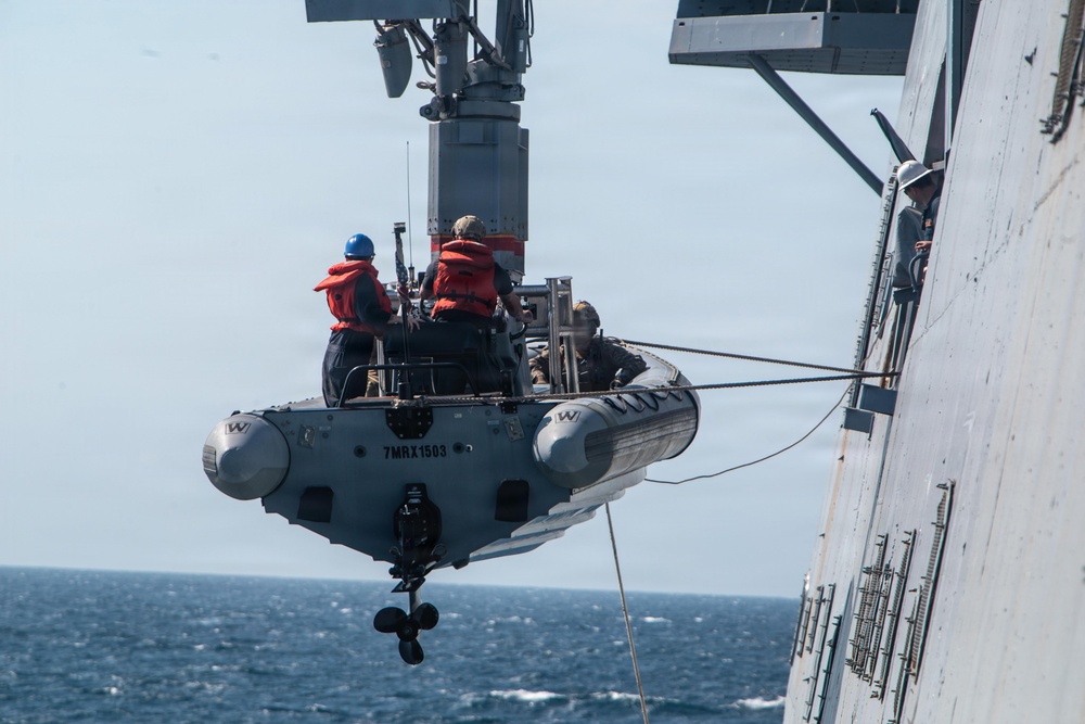 A rigid-hull inflatable boat (RHIB) is being hoisted or lowered by a davit crane alongside a naval warship at sea. At least three personnel are visible aboard the RHIB, wearing orange life vests and a blue hard hat, while additional crew members observe from the ship's side. The boat bears the identifier '7MRX1503' on its hull.