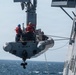 A rigid-hull inflatable boat (RHIB) is being hoisted or lowered by a davit crane alongside a naval warship at sea. At least three personnel are visible aboard the RHIB, wearing orange life vests and a blue hard hat, while additional crew members observe from the ship's side. The boat bears the identifier '7MRX1503' on its hull.