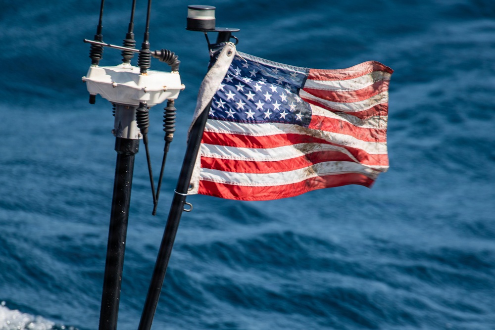 A weathered and worn American flag is mounted on a pole at the stern or mast of a vessel, flying in the wind over open blue ocean water. The flag shows visible signs of age and use, including discoloration, staining, and fraying. Navigation or communication equipment is visible on adjacent mast hardware.
