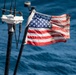 A weathered and worn American flag is mounted on a pole at the stern or mast of a vessel, flying in the wind over open blue ocean water. The flag shows visible signs of age and use, including discoloration, staining, and fraying. Navigation or communication equipment is visible on adjacent mast hardware.