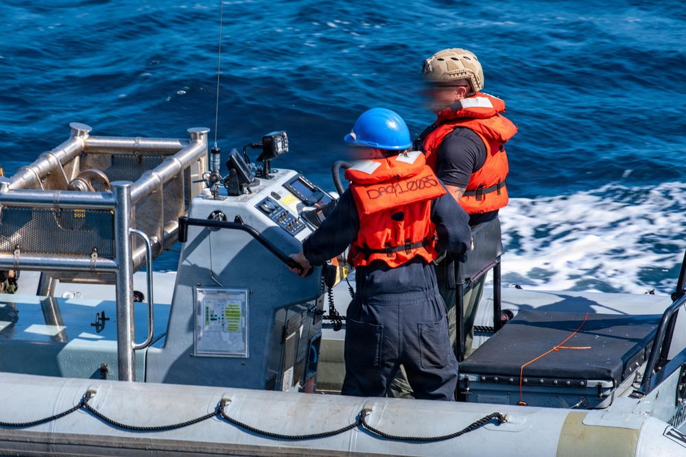 Two personnel are operating a rigid-hull inflatable boat (RHIB) underway on open blue water. Both individuals are wearing orange life vests and are positioned at the helm console, which features navigation and communication equipment. The boat appears to be moving at speed, evidenced by the white wake visible alongside the hull.
