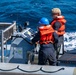 Two personnel are operating a rigid-hull inflatable boat (RHIB) underway on open blue water. Both individuals are wearing orange life vests and are positioned at the helm console, which features navigation and communication equipment. The boat appears to be moving at speed, evidenced by the white wake visible alongside the hull.