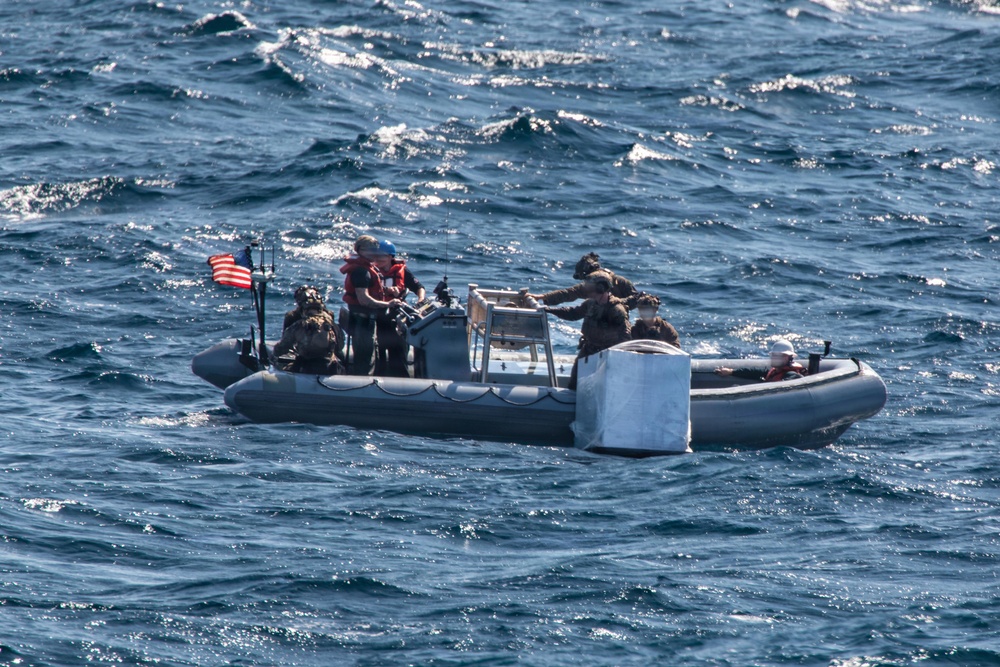 A gray rigid inflatable boat (RIB) operated by what appears to be U.S. military or naval personnel is underway on open ocean waters. The crew, wearing tactical gear and helmets, appears to be recovering or securing a large white object at the bow of the vessel. An American flag is visibly mounted on the boat.