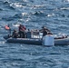 A gray rigid inflatable boat (RIB) operated by what appears to be U.S. military or naval personnel is underway on open ocean waters. The crew, wearing tactical gear and helmets, appears to be recovering or securing a large white object at the bow of the vessel. An American flag is visibly mounted on the boat.