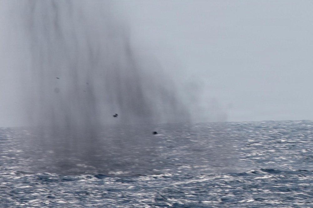 The image shows a large plume of mist or spray rising from the ocean surface, consistent with a whale blow or spout. The scene is set over choppy open ocean water under an overcast, hazy sky. Two small dark birds are visible flying near the base of the spray column.