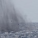 The image shows a large plume of mist or spray rising from the ocean surface, consistent with a whale blow or spout. The scene is set over choppy open ocean water under an overcast, hazy sky. Two small dark birds are visible flying near the base of the spray column.