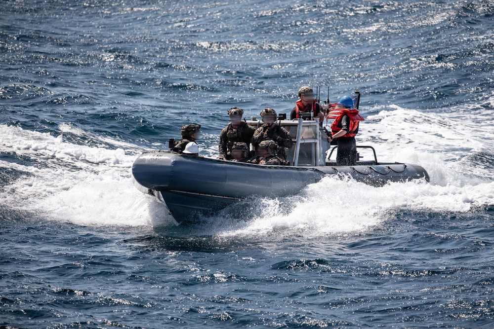 A rigid-hull inflatable boat (RHIB) is underway at speed on open blue water, carrying several personnel in military tactical gear along with two individuals in civilian safety equipment. The boat is generating significant wake and spray, indicating high-speed travel. Faces of military personnel have been obscured, suggesting operational security considerations.