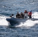 A rigid-hull inflatable boat (RHIB) is underway at speed on open blue water, carrying several personnel in military tactical gear along with two individuals in civilian safety equipment. The boat is generating significant wake and spray, indicating high-speed travel. Faces of military personnel have been obscured, suggesting operational security considerations.