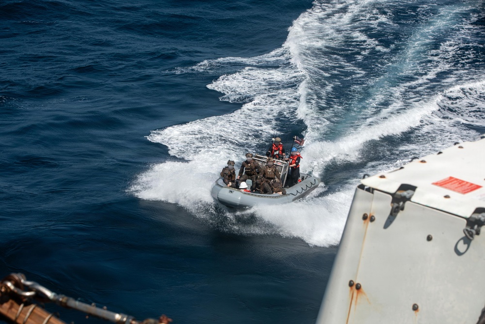 An aerial view of a rigid-hull inflatable boat (RHIB) operating at high speed on open ocean, viewed from the deck of a larger vessel. The RHIB carries multiple personnel in tactical/military gear, suggesting a military or law enforcement boarding operation. The image is taken from an elevated position on the parent vessel, with wake turbulence visible behind the small craft.