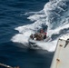 An aerial view of a rigid-hull inflatable boat (RHIB) operating at high speed on open ocean, viewed from the deck of a larger vessel. The RHIB carries multiple personnel in tactical/military gear, suggesting a military or law enforcement boarding operation. The image is taken from an elevated position on the parent vessel, with wake turbulence visible behind the small craft.