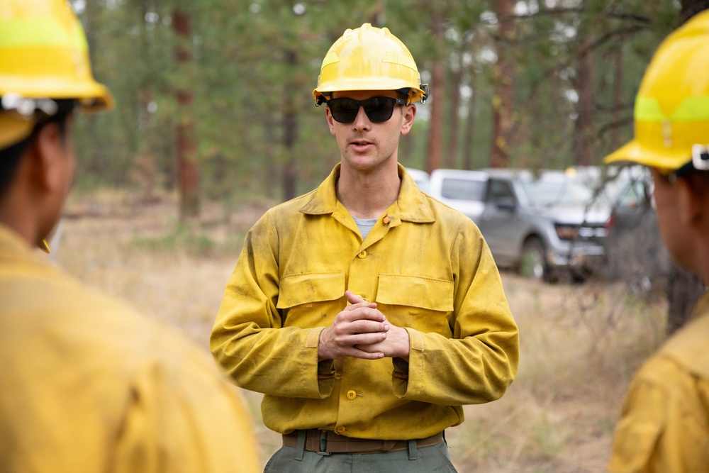 A wildland firefighter in standard Nomex gear addresses a small group of colleagues in a forested setting. The central figure is wearing a yellow hard hat, yellow fire-resistant shirt, and dark sunglasses, with hands clasped in a briefing posture. The scene appears to be a pre- or post-operation crew briefing near parked vehicles in a pine forest.