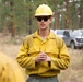 A wildland firefighter in standard Nomex gear addresses a small group of colleagues in a forested setting. The central figure is wearing a yellow hard hat, yellow fire-resistant shirt, and dark sunglasses, with hands clasped in a briefing posture. The scene appears to be a pre- or post-operation crew briefing near parked vehicles in a pine forest.