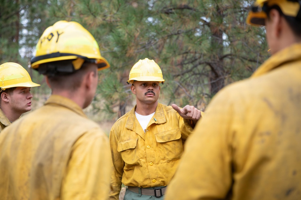 A group of wildland firefighters in yellow Nomex fire-resistant gear and yellow hard hats are gathered in a forested area for what appears to be a briefing or debriefing. The central figure, facing the camera, is gesturing with his finger as if giving instructions or making a point to the others. The setting among pine trees and the worn, dirty condition of their gear suggests active field operations.
