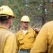 A group of wildland firefighters in yellow Nomex fire-resistant gear and yellow hard hats are gathered in a forested area for what appears to be a briefing or debriefing. The central figure, facing the camera, is gesturing with his finger as if giving instructions or making a point to the others. The setting among pine trees and the worn, dirty condition of their gear suggests active field operations.