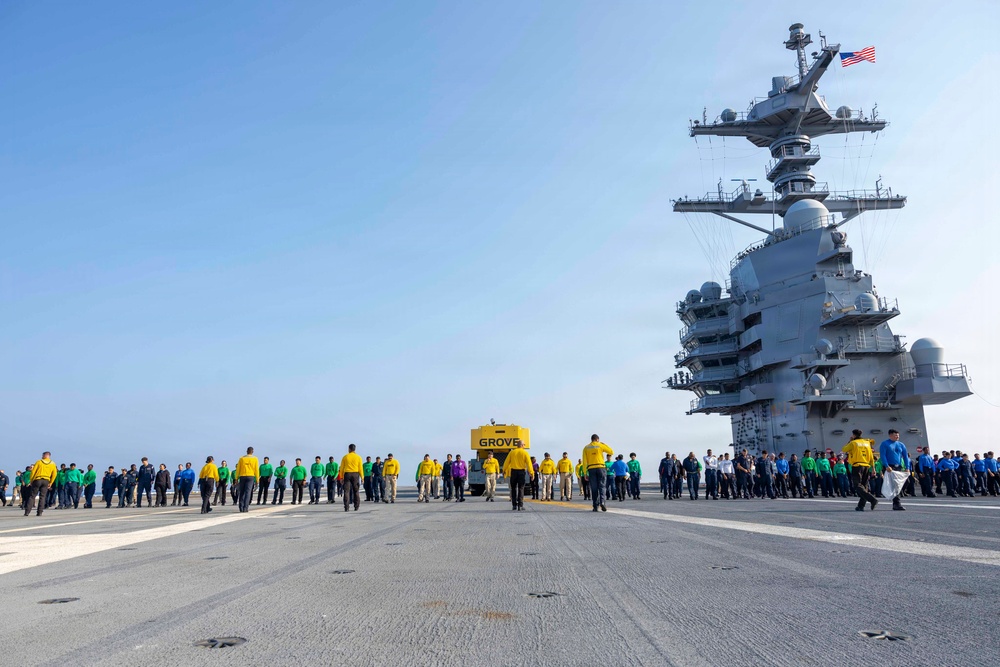 A large group of U.S. Navy personnel are conducting a Foreign Object Debris (FOD) walkdown on the flight deck of an aircraft carrier. Crew members wearing color-coded jerseys — yellow, green, and blue — are walking in a line formation across the deck, searching for debris. The ship's island superstructure is visible on the right side, flying an American flag.