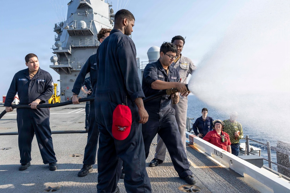U.S. Navy sailors are conducting a firefighting drill on the deck of a naval vessel, likely an aircraft carrier. Personnel in Navy coveralls and working uniforms are operating a fire hose, directing a water stream over the ship's side. The exercise appears to be a damage control training evolution with multiple crew members participating in various roles.