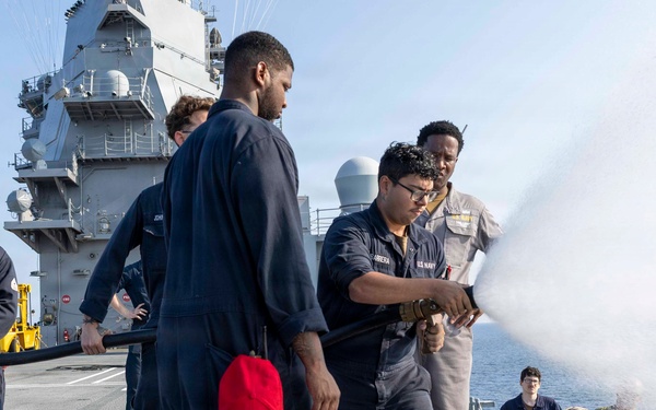 U.S. Navy sailors are conducting a firefighting drill on the deck of a naval vessel, likely an aircraft carrier. Personnel in Navy coveralls and working uniforms are operating a fire hose, directing a water stream over the ship's side. The exercise appears to be a damage control training evolution with multiple crew members participating in various roles.