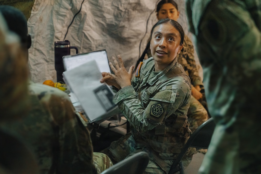 A female soldier in US Army OCP (Operational Camouflage Pattern) uniform is actively briefing or presenting to a group inside what appears to be a military field tent or temporary structure. She is holding a binder or document folder and gesturing expressively while speaking. Additional soldiers in uniform are visible in the background, suggesting a group training or planning session.