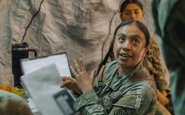 A female soldier in US Army OCP (Operational Camouflage Pattern) uniform is actively briefing or presenting to a group inside what appears to be a military field tent or temporary structure. She is holding a binder or document folder and gesturing expressively while speaking. Additional soldiers in uniform are visible in the background, suggesting a group training or planning session.
