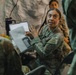 A female soldier in US Army OCP (Operational Camouflage Pattern) uniform is actively briefing or presenting to a group inside what appears to be a military field tent or temporary structure. She is holding a binder or document folder and gesturing expressively while speaking. Additional soldiers in uniform are visible in the background, suggesting a group training or planning session.