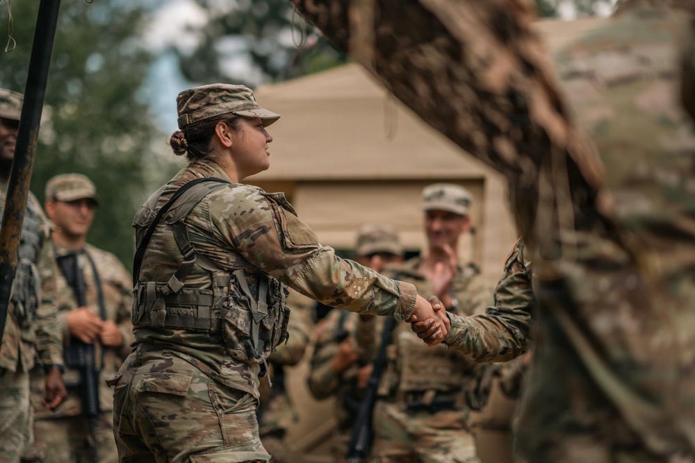 A female soldier in MultiCam/OCP camouflage uniform and tactical vest is shaking hands with another soldier, partially visible on the right edge of the frame. The scene appears to take place at a military field camp or training area, with a tan military tent visible in the background. Several other soldiers in similar uniforms are visible in the background, some smiling and observing the interaction.
