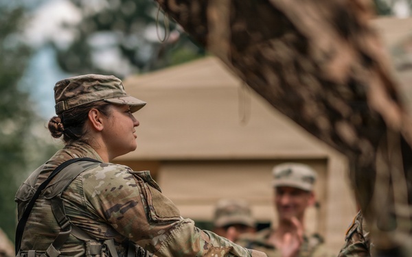 A female soldier in MultiCam/OCP camouflage uniform and tactical vest is shaking hands with another soldier, partially visible on the right edge of the frame. The scene appears to take place at a military field camp or training area, with a tan military tent visible in the background. Several other soldiers in similar uniforms are visible in the background, some smiling and observing the interaction.