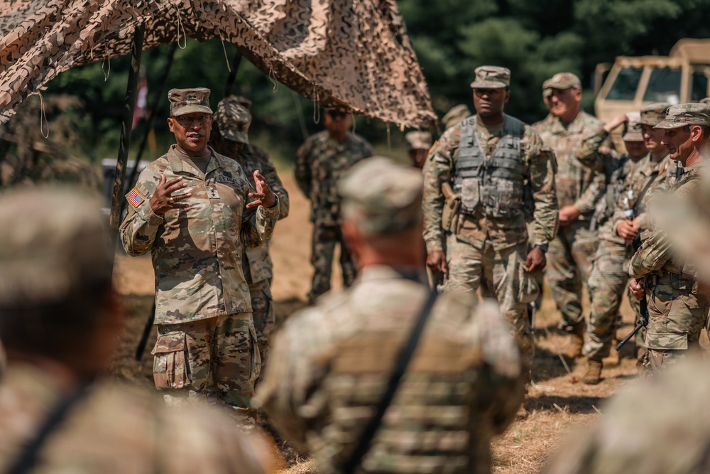 A U.S. Army officer, visibly wearing general-grade rank insignia and an American flag patch, addresses a group of soldiers in an outdoor field setting beneath camouflage netting. Multiple soldiers in OCP (Operational Camouflage Pattern) uniforms are gathered, some wearing tactical vests, suggesting a briefing or after-action review during a training exercise or field operation. Military vehicles are partially visible in the background.