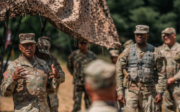 A U.S. Army officer, visibly wearing general-grade rank insignia and an American flag patch, addresses a group of soldiers in an outdoor field setting beneath camouflage netting. Multiple soldiers in OCP (Operational Camouflage Pattern) uniforms are gathered, some wearing tactical vests, suggesting a briefing or after-action review during a training exercise or field operation. Military vehicles are partially visible in the background.