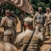 A U.S. Army officer, visibly wearing general-grade rank insignia and an American flag patch, addresses a group of soldiers in an outdoor field setting beneath camouflage netting. Multiple soldiers in OCP (Operational Camouflage Pattern) uniforms are gathered, some wearing tactical vests, suggesting a briefing or after-action review during a training exercise or field operation. Military vehicles are partially visible in the background.