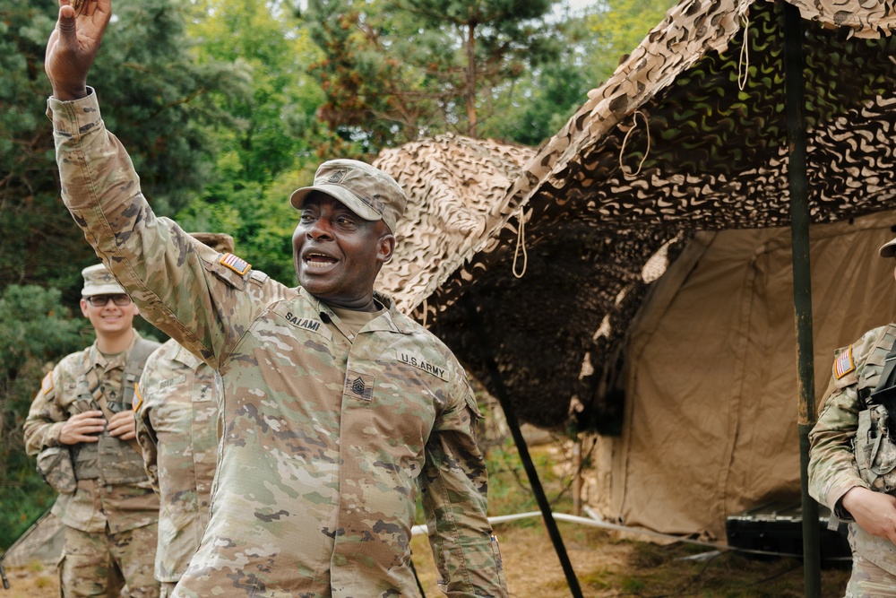 A U.S. Army soldier with the name tape 'SALAMI' and a Command Sergeant Major rank insignia is gesturing with his right arm raised, appearing to be speaking or giving instructions in a field environment. He is surrounded by other soldiers in OCP (Operational Camouflage Pattern) uniforms near a military field tent covered with camouflage netting. The setting appears to be an outdoor military training or operational area with trees in the background.