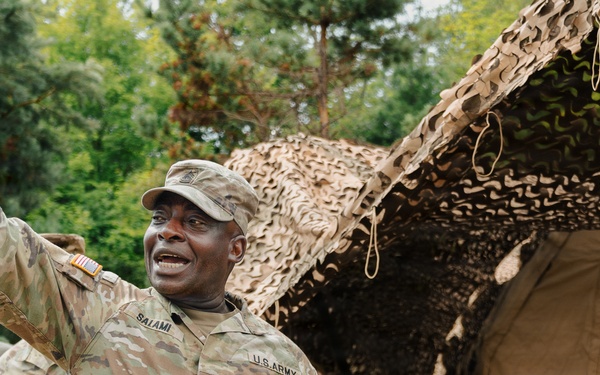 A U.S. Army soldier with the name tape 'SALAMI' and a Command Sergeant Major rank insignia is gesturing with his right arm raised, appearing to be speaking or giving instructions in a field environment. He is surrounded by other soldiers in OCP (Operational Camouflage Pattern) uniforms near a military field tent covered with camouflage netting. The setting appears to be an outdoor military training or operational area with trees in the background.